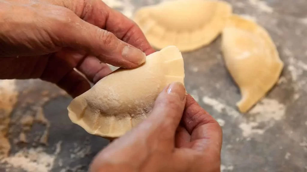  Hände formen eine ungekochte Pirogge auf einer bemehlten Arbeitsfläche.